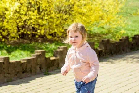 Cheerful little girl with blond hair running near blooming yellow forsythia bush